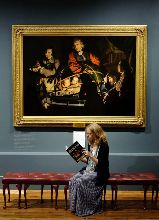 A woman reading a book in front of a painting in an art gallery.