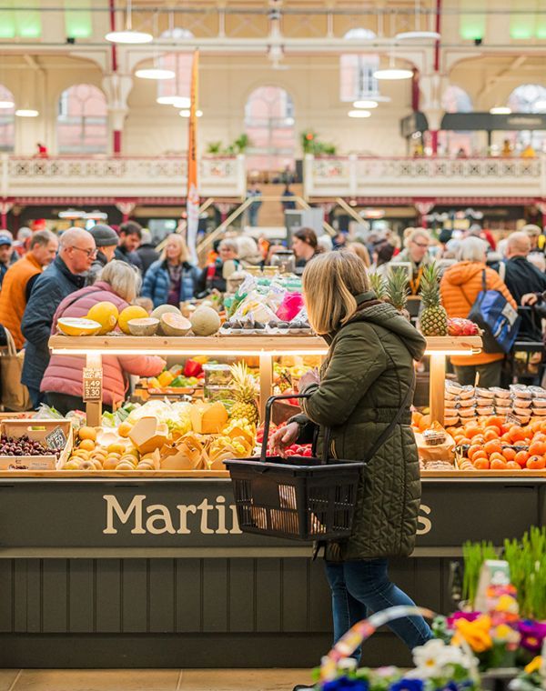 A woman carrying a shopping basket selects produce at the busy Farmer's Market, with various fruits and vegetables displayed on the stall.